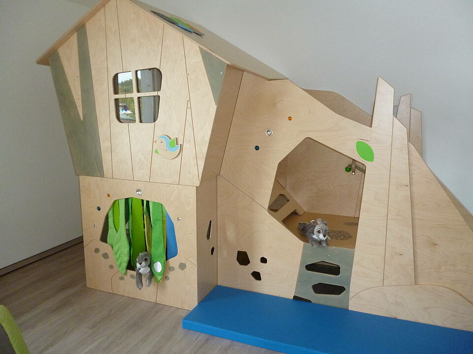 Kletterlandschaft gestaltet wie ein Haus mit Tür und Fenster. Davor liegt eine Matte