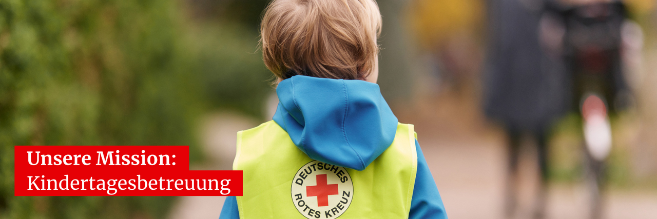 Kind steht mit dem Rücken zum Betrachter. Kind hat eine Warnweste mit dem DRK-Logo an und befindet sich draußen