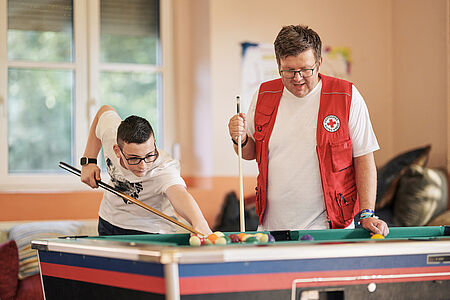 Ein Mitarbeitender des DRK mit DRK-Kleidung spielt mit einem Jugendlichen Billiard